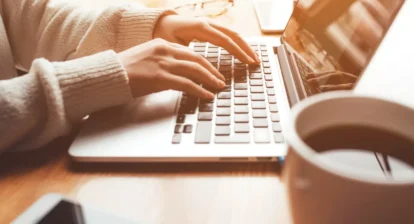 The picture shows a person sitting at a desk and typing on a laptop. The person is wearing a white sweater and the laptop is open. There is a cup of coffee on the desk to the right of the laptop. The person is probably working on something or browsing the internet.
